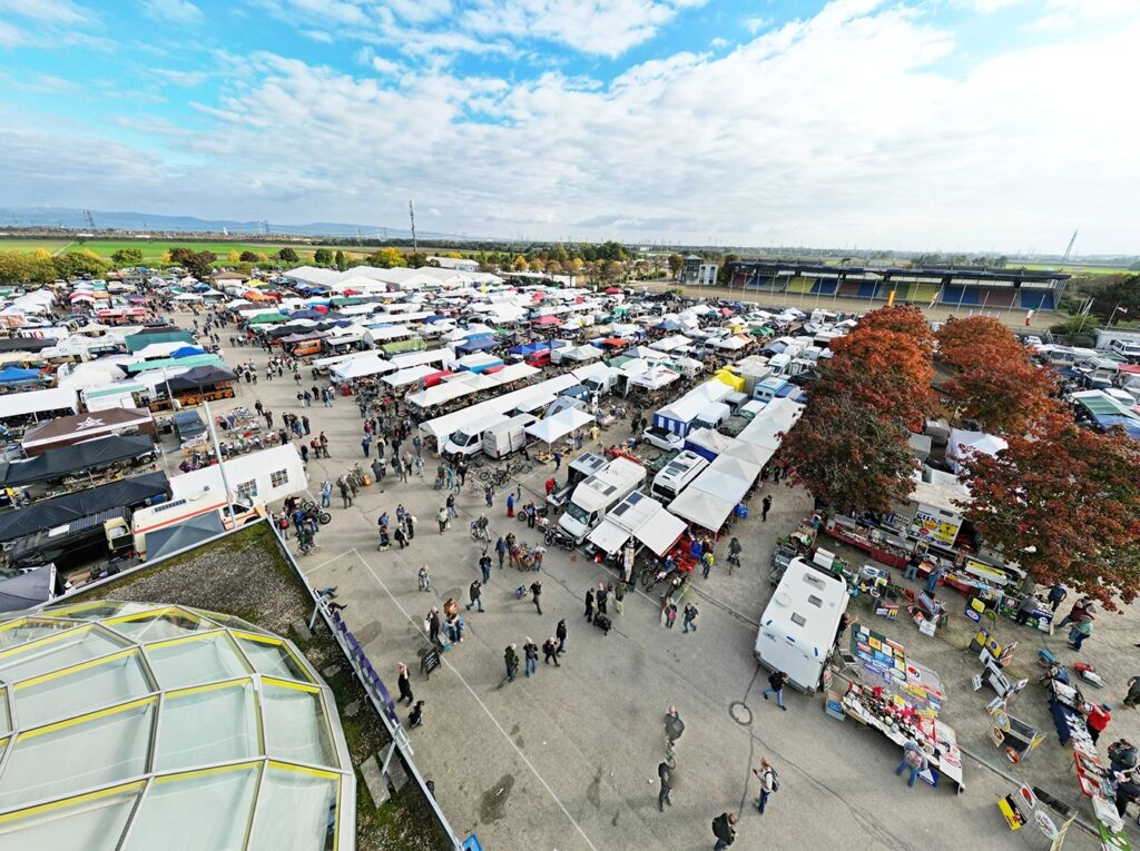 Luftaufnahme eines großen Freiluftmarkts mit Reihen von Marktzelten, Verkaufsständen, Menschen und parkenden Fahrzeugen, herbstliche Bäume im Bild