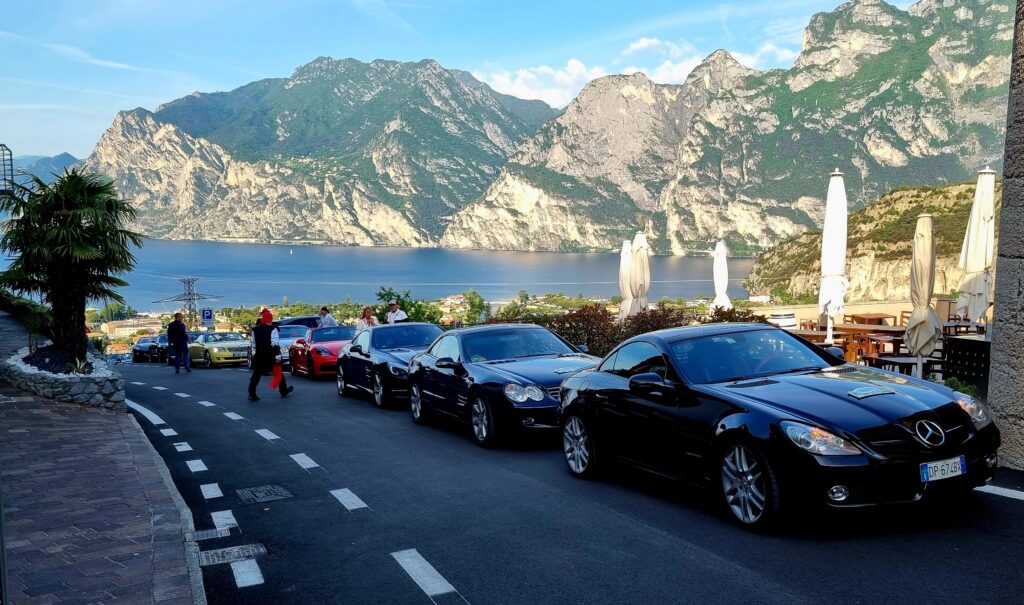 Mehrere Sportwagen an einer Küstenstraße mit Blick auf einen See und hohe Berge, einige Passanten und Außenbestuhlung am Straßenrand.