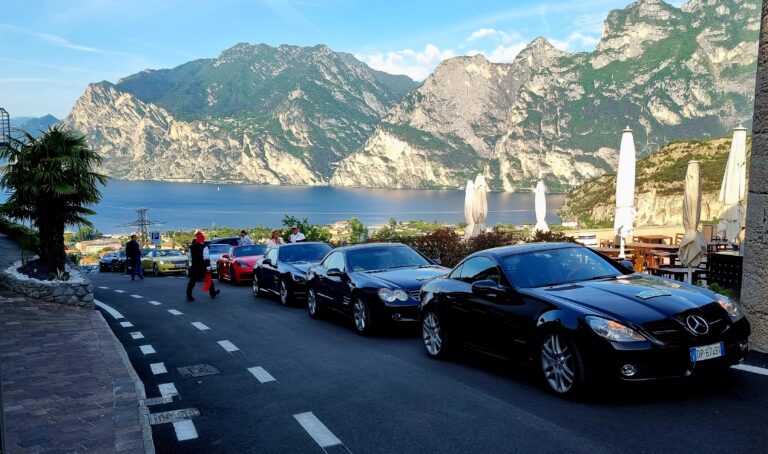 Mehrere Sportwagen an einer Küstenstraße mit Blick auf einen See und hohe Berge, einige Passanten und Außenbestuhlung am Straßenrand.