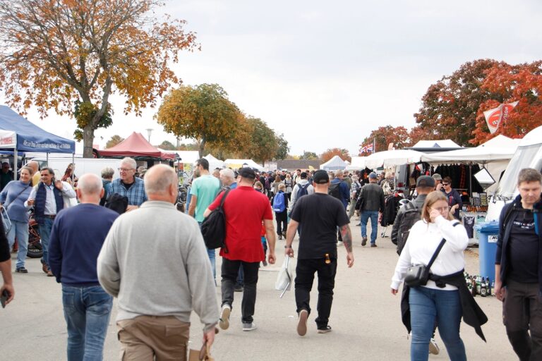 Freiluftmarkt mit vielen Menschen, Verkaufsständen und bunten Herbstbäumen im Hintergrund.