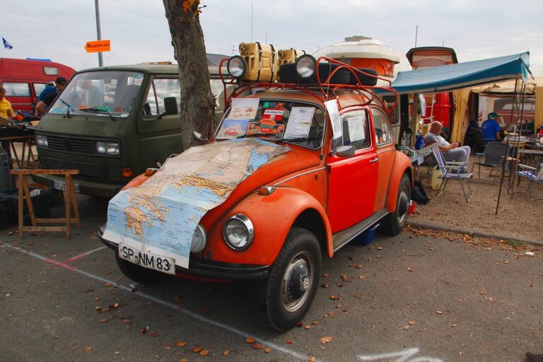 Roter VW-Käfer mit aufliegender Landkarte, Dachgepäck und Marktständen sowie Personen im Hintergrund.