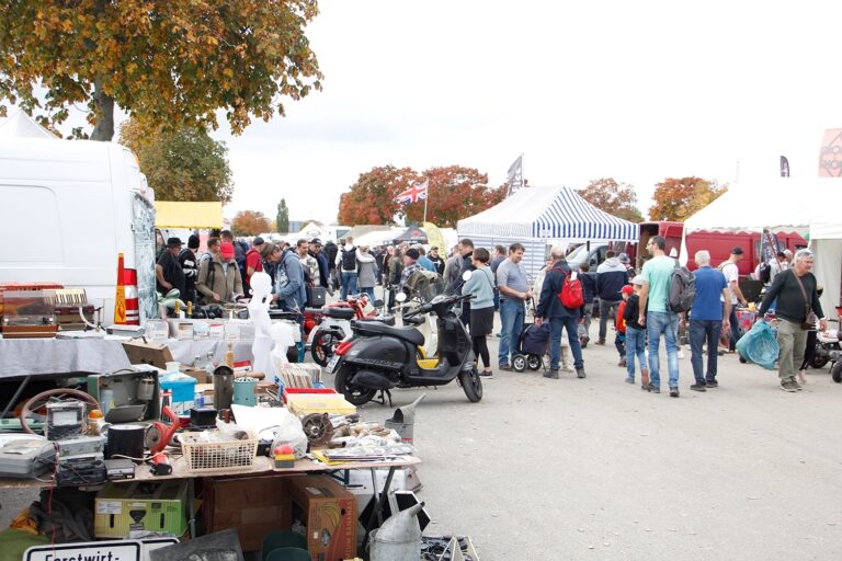 Freiluft-Flohmarkt mit Marktständen, Menschenmengen und mehreren Rollern im Vordergrund.
