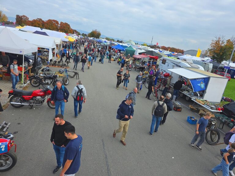 Blick auf einen belebten Flohmarkt im Freien mit Verkaufszelten, Menschen und mehreren Motorrädern.