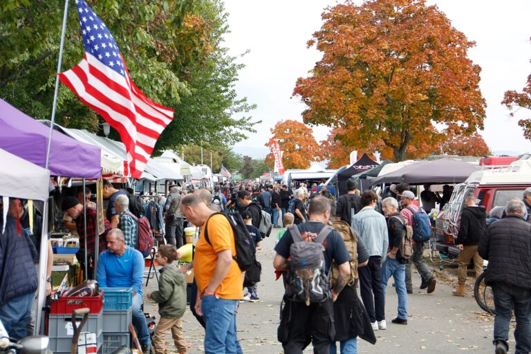 Belebter Straßenmarkt mit vielen Besuchern und Marktständen; eine US-Flagge hängt links, im Hintergrund Bäume mit herbstlichem Laub.