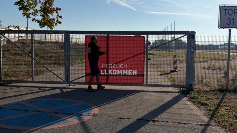 Person vor verschlossenem Gittertor mit rotem Schild 'Herzlich Willkommen' auf einem Freigelände bei Sonnenschein.