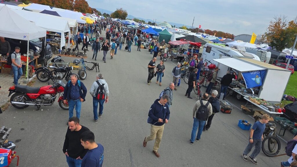 Blick von oben auf einen belebten Außenmarkt mit Reihen von Ständen, ausgestellten Motorrädern und Menschen, die über den Platz gehen.