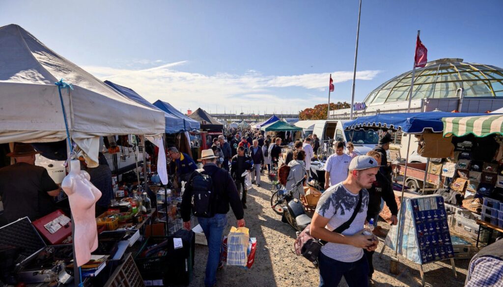 Blick über einen belebten Flohmarkt mit Verkaufsständen, Auslageartikeln und vielen Menschen an einem sonnigen Tag.