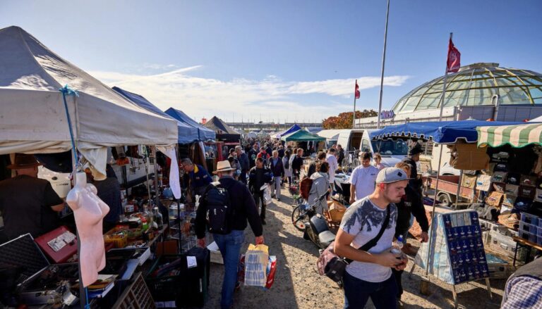 Blick über einen belebten Flohmarkt mit Verkaufsständen, Auslageartikeln und vielen Menschen an einem sonnigen Tag.
